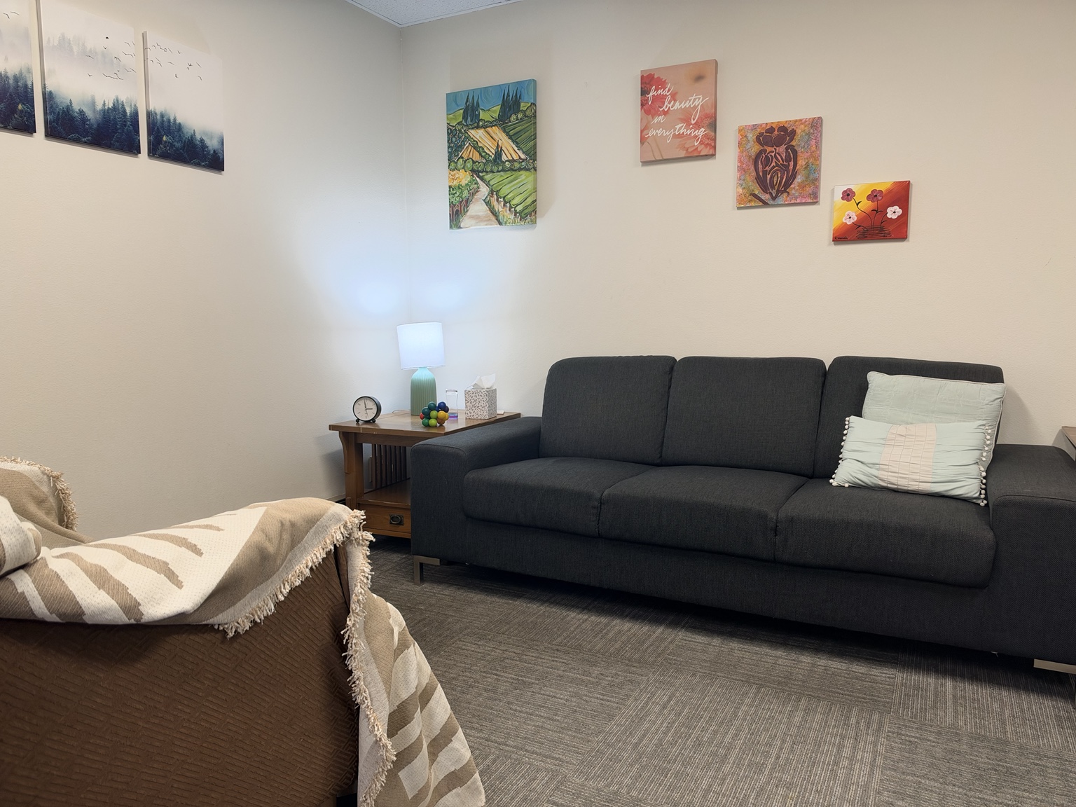 Storyboard Therapy office with a charcoal couch, soft pillows, a blanket-covered armchair, forest and owl artwork, and a side table with a lamp, tissues, and a fidget toy.